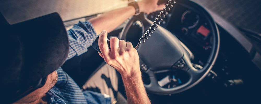 Looking down at a trucker wearing a dark blue baseball hat. He's driving, with one hand on the steering wheel and the other is holding up a radio to speak into.